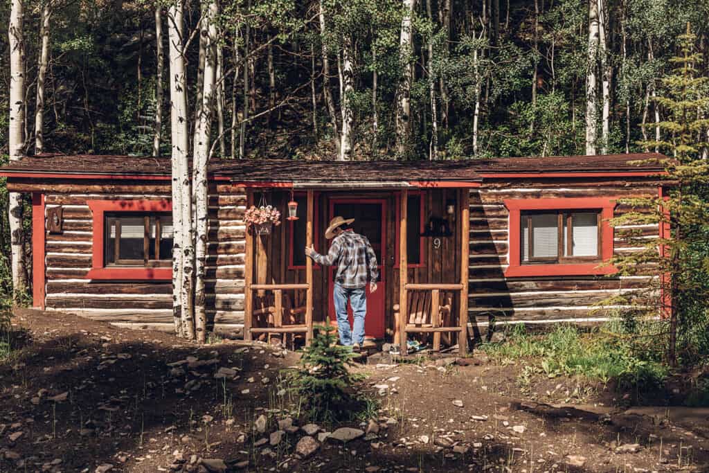 A man standing in front of a log cabin in the woods.