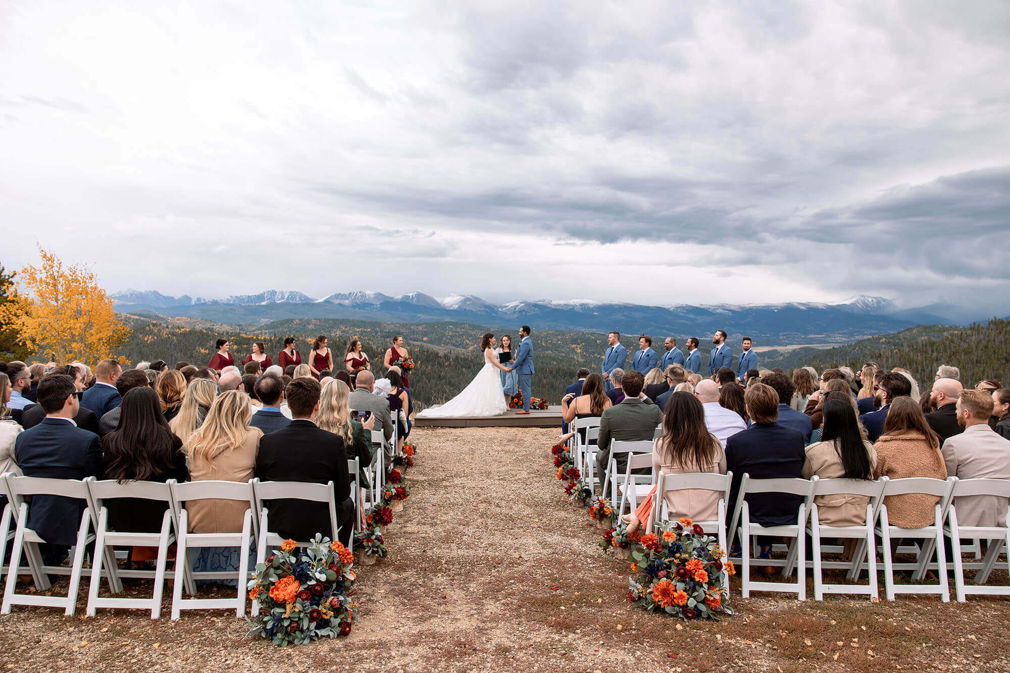 A couple exchanges vows on a mountain top with a breathtaking view of the Rocky Mountains in the background, surrounded by their wedding party and guests seated on white chairs at Granby Ranch in Grand County Colorado.