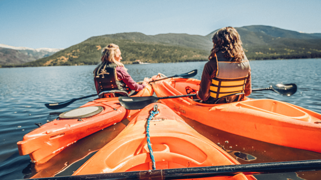 Two women in orange kayaks paddle across Grand Lake with mountains in the background.