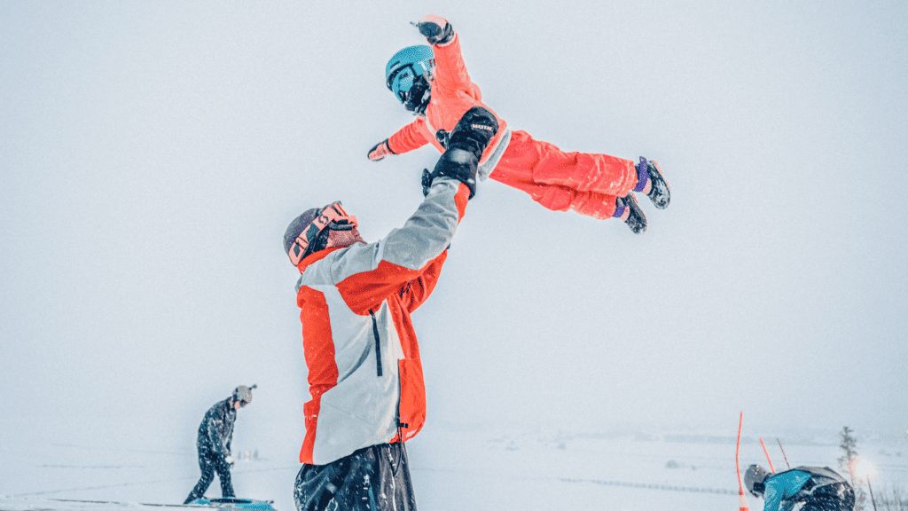 A person holds a child up in the air during a snow storm.  Their bright pink and red clothing contrasts with the falling snow.