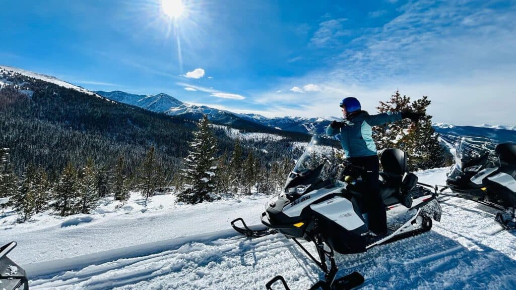 Two people on snowmobiles in Grand County. They take a break on the trail while the sun beats down on them from a bright blue sky.