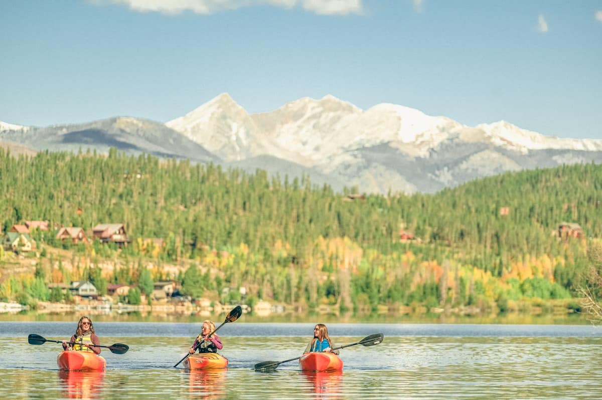 Three people kayak on Grand Lake with snowcapped Rocky Mountain National Park peaks and golden aspens in the background.