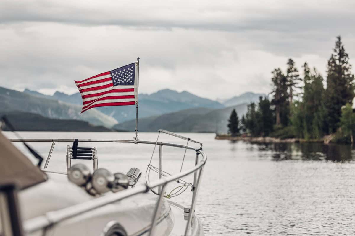 A motor boat moves on Lake Granby, with an American flag at the bow. Mountains and trees can be seen along the shoreline