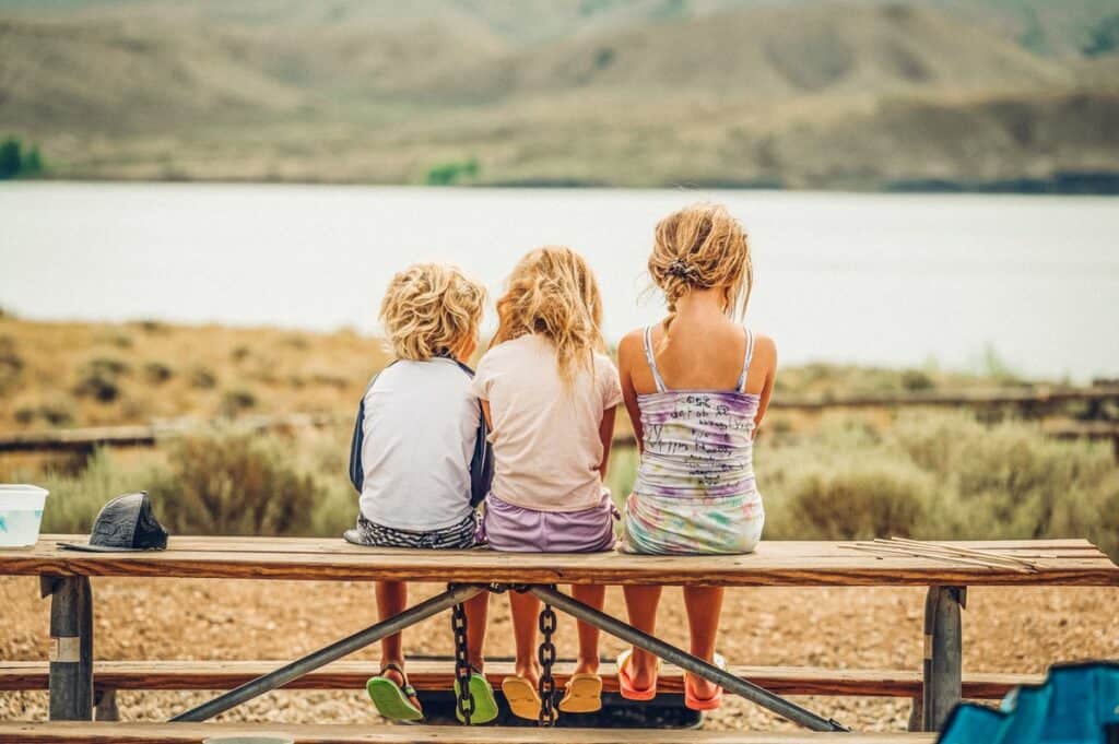 Three children sitting on a bench overlooking Lake Granby facing away from the camera, with scenic hills in the background.