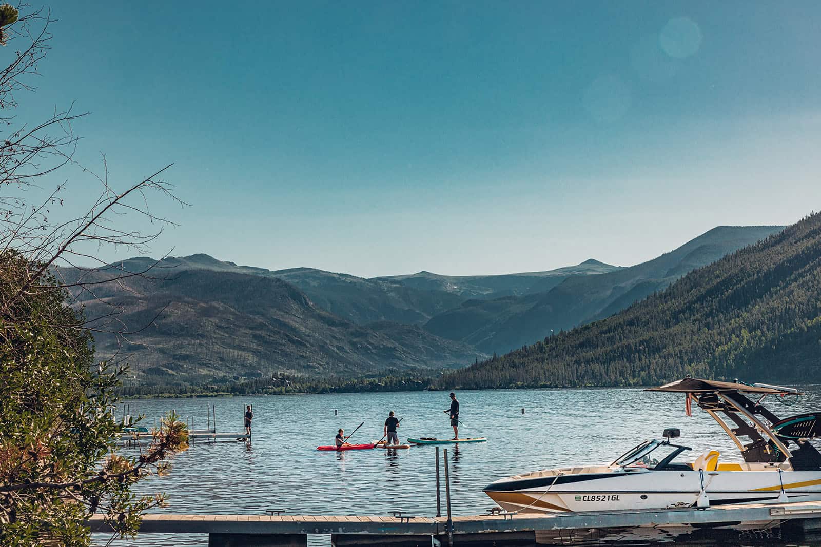 A serene lake scene with people paddleboarding near a dock with a motorboat and mountains in the background on a sunny day.
