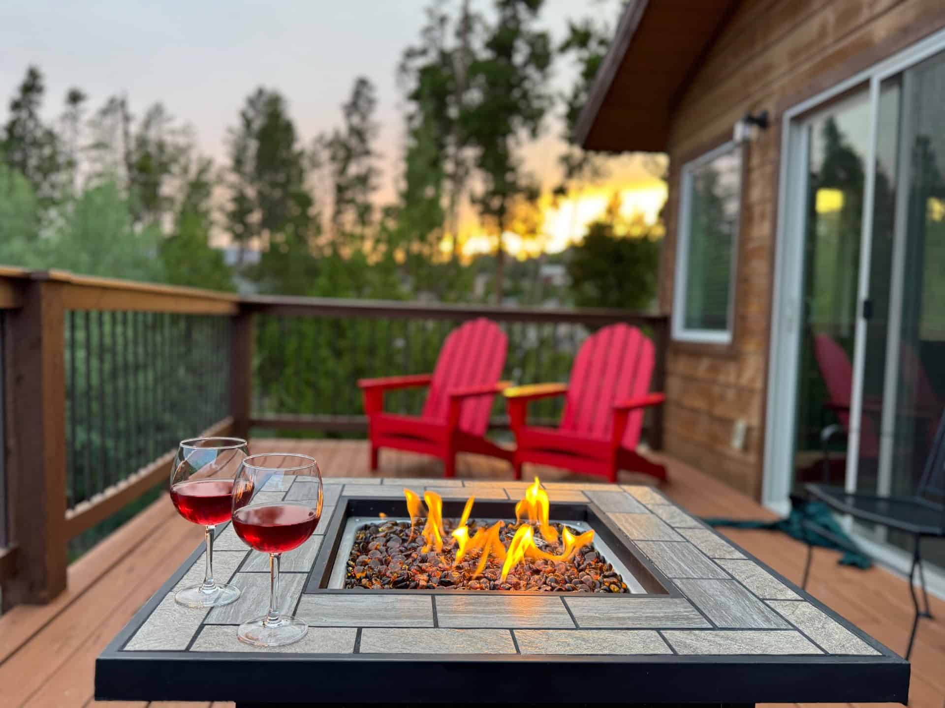 A cozy deck with a fire pit table, red Adirondack chairs, and wine glasses overlooking a forest at sunset in Grand County Colorado.