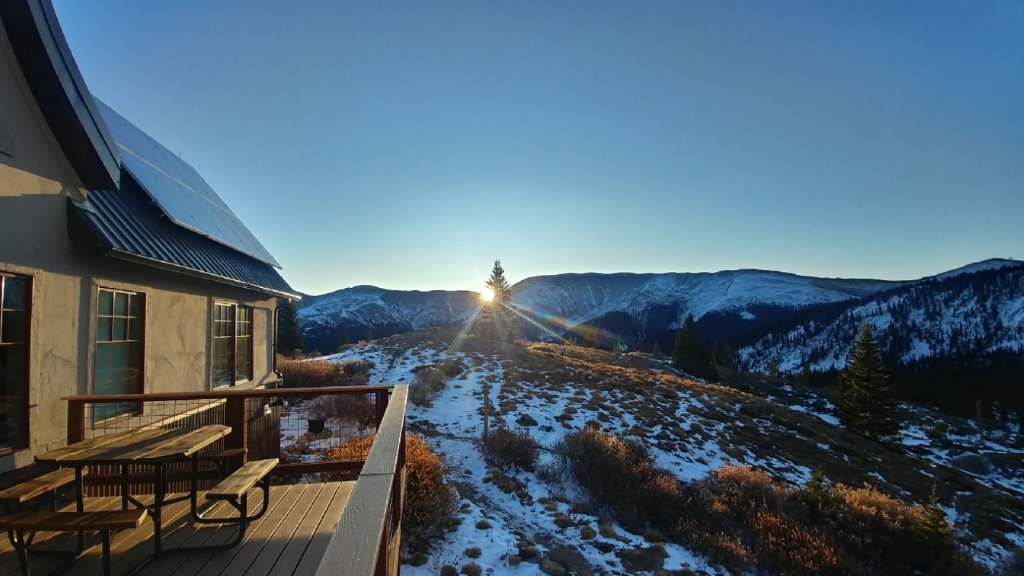 Sunrise over snow-capped mountains with a wooden cabin and picnic table in the foreground in Grand County Colorado.