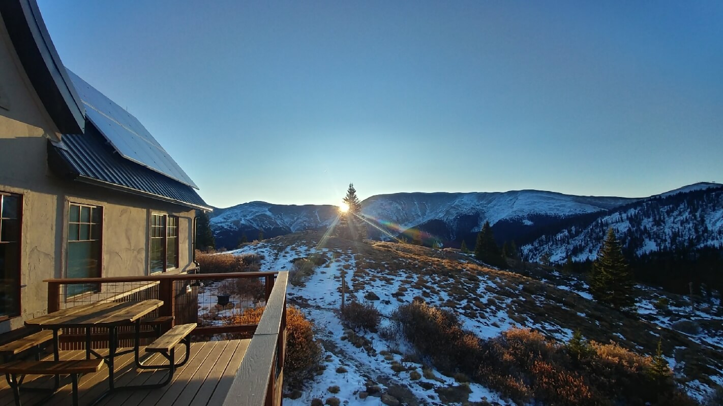 Sunrise over snow-capped mountains with a wooden cabin and picnic table in the foreground in Grand County Colorado.