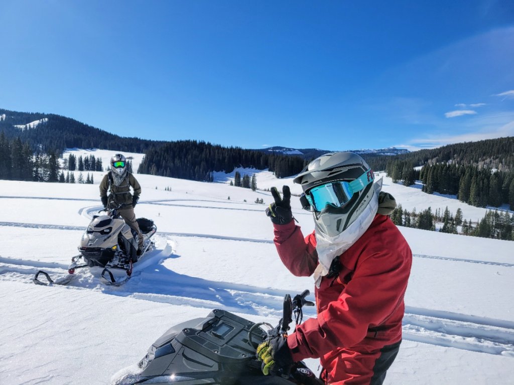 Two snowmobilers in winter gear ride across a snowy landscape with mountains in the background in Grand County Colorado.