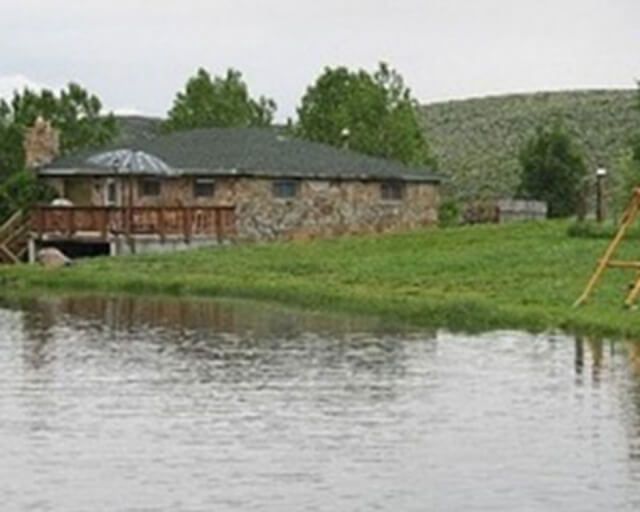 A rustic stone house with a deck overlooks a serene lake surrounded by lush greenery in Grand County Colorado.