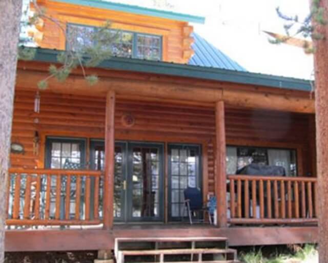 A rustic log cabin with a spacious porch and large windows overlooks a forested area in Grand County Colorado.