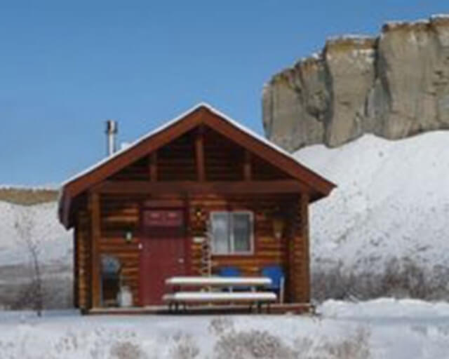 A rustic log cabin with a red door sits in a snowy landscape near a rocky cliff in Grand County Colorado.