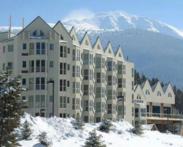 A multi-story mountain lodge with snow-covered trees and a majestic peak in the background in Grand County Colorado.