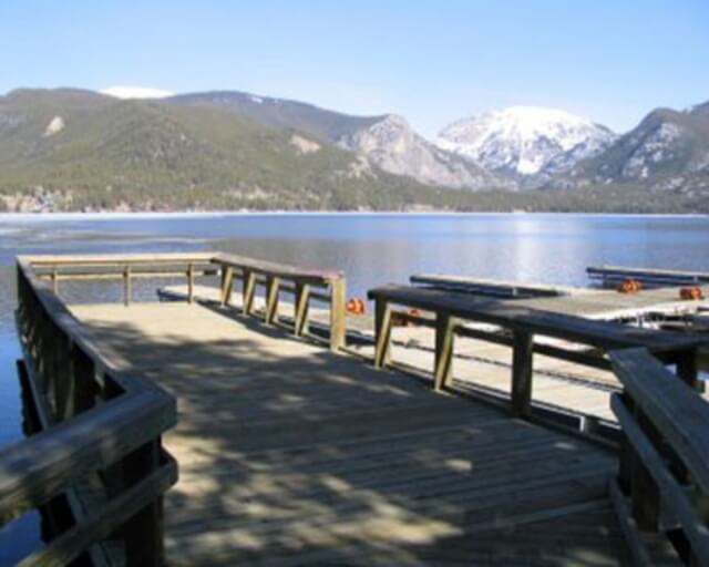 A wooden dock extends over a calm lake with snow-capped mountains in the distance in Grand County Colorado.
