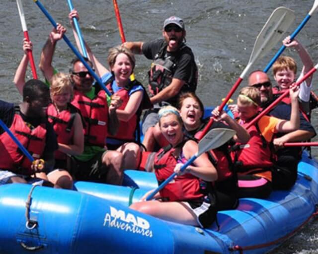 A group of people in life jackets paddle a blue raft down a river in Grand County Colorado.