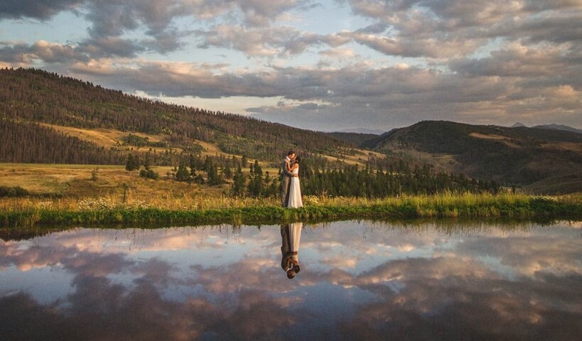 A couple embraces at sunset by a serene pond reflecting mountains and clouds in Grand County Colorado.