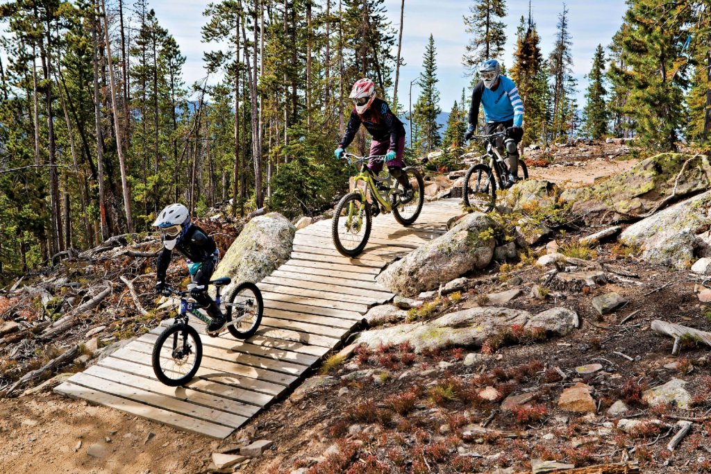 Three mountain bikers, including a child, ride down a wooden ramp in a forest in Grand County Colorado.