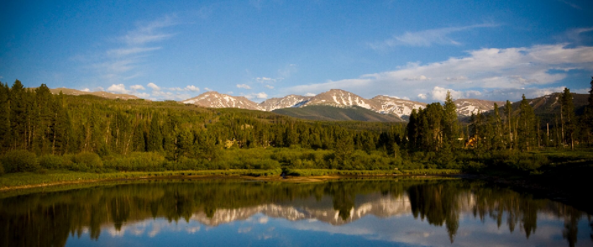 A serene mountain lake reflects snow-capped peaks and evergreen forests in Grand County Colorado.