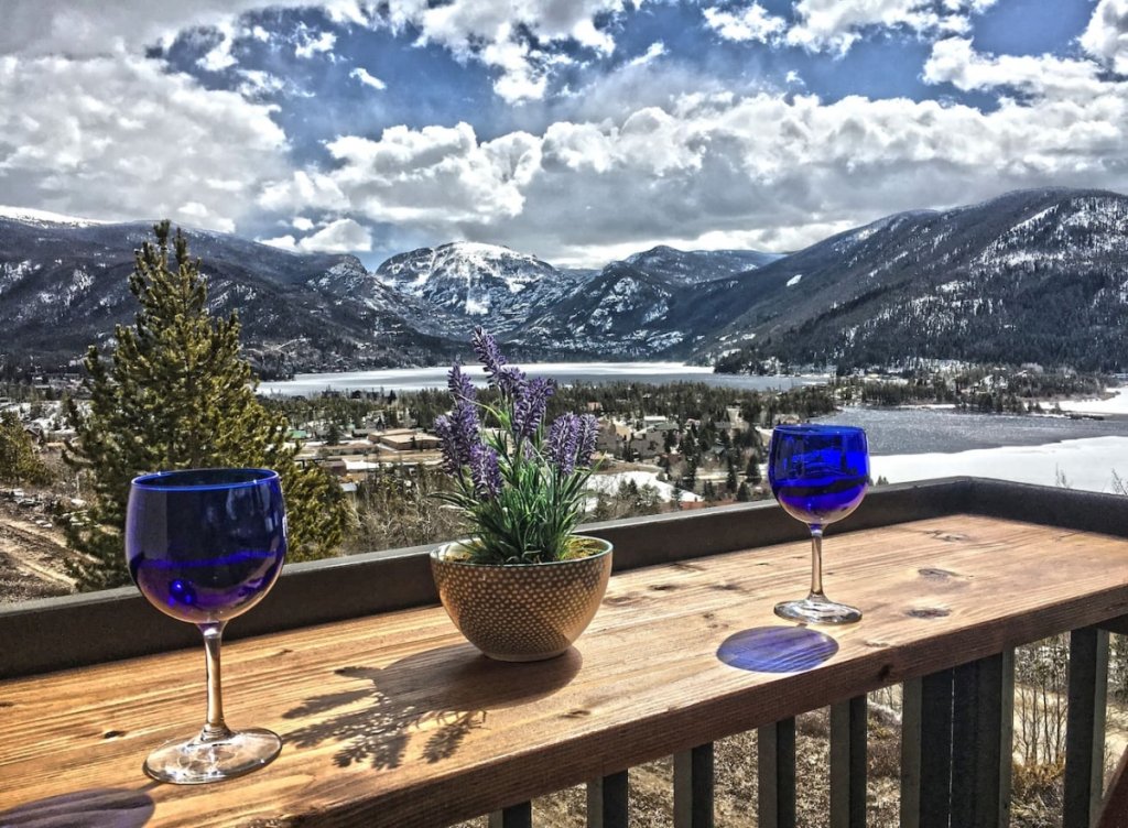 A wooden deck with blue wine glasses and a lavender plant overlooks a snowy mountain range and lake in Grand County Colorado.