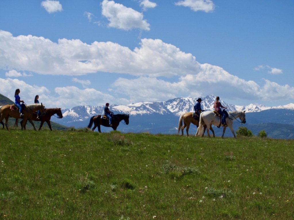 Five horseback riders traverse a grassy field with snow-capped mountains in the distance in Grand County Colorado.