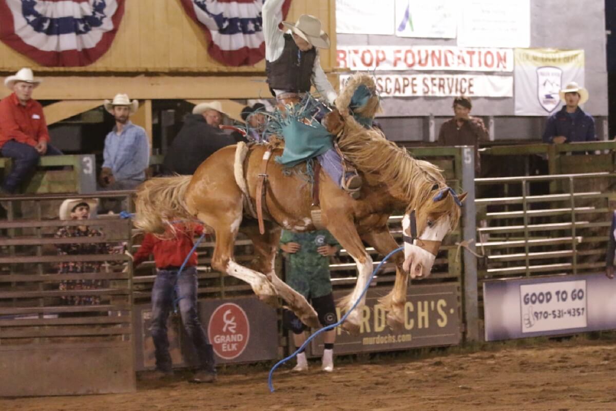 Cowboy riding bucking bronco at rodeo, waving American flag in victory in Grand County Colorado.