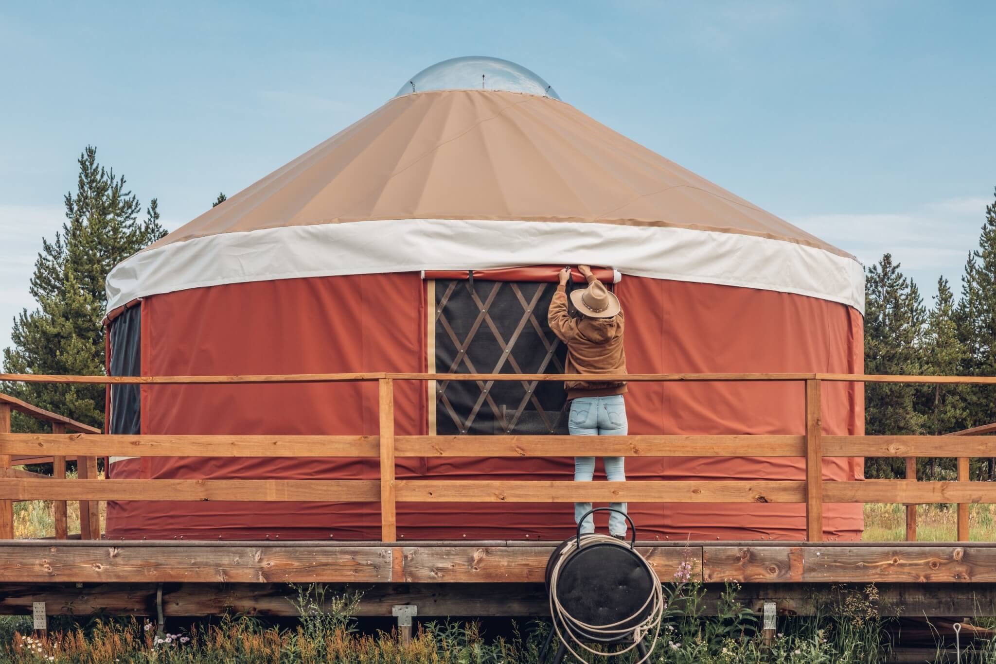 A person wearing a hat adjusts the entrance of a large, round, beige and red tent with a wooden deck in Grand County Colorado.