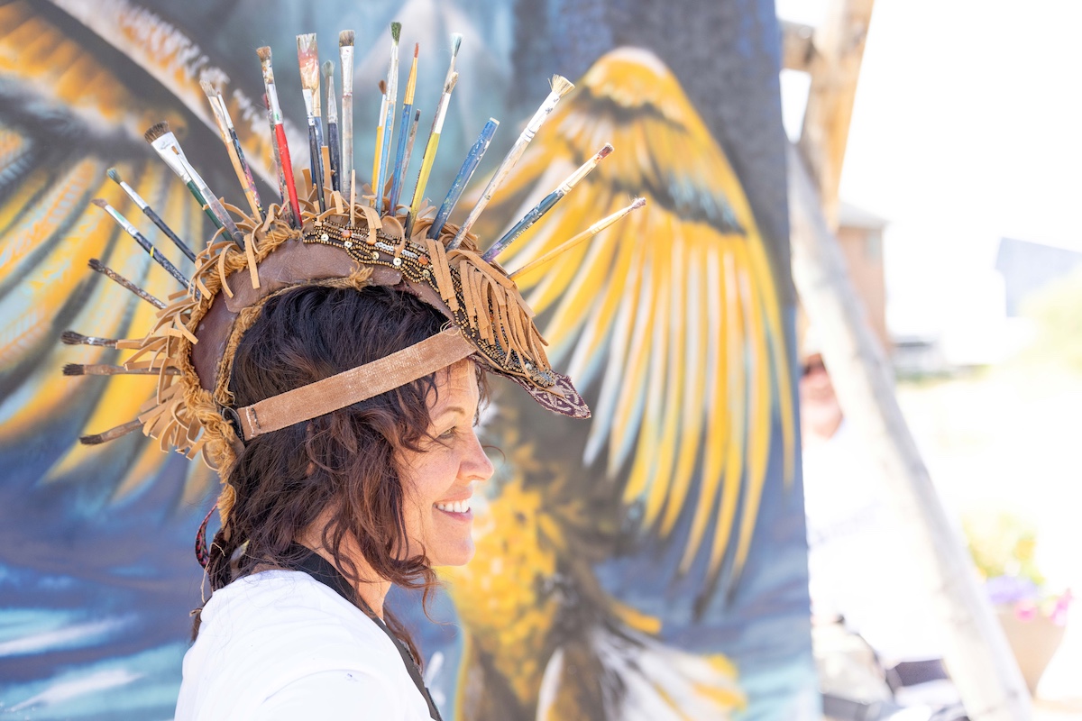 A woman with a crown of paintbrushes stands in front of a mural of a bird with yellow wings in Grand County Colorado.