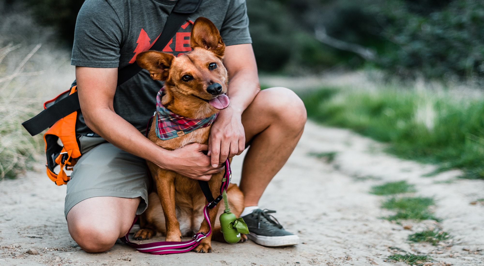 Person kneeling on a trail with a happy dog wearing a bandana and leash.