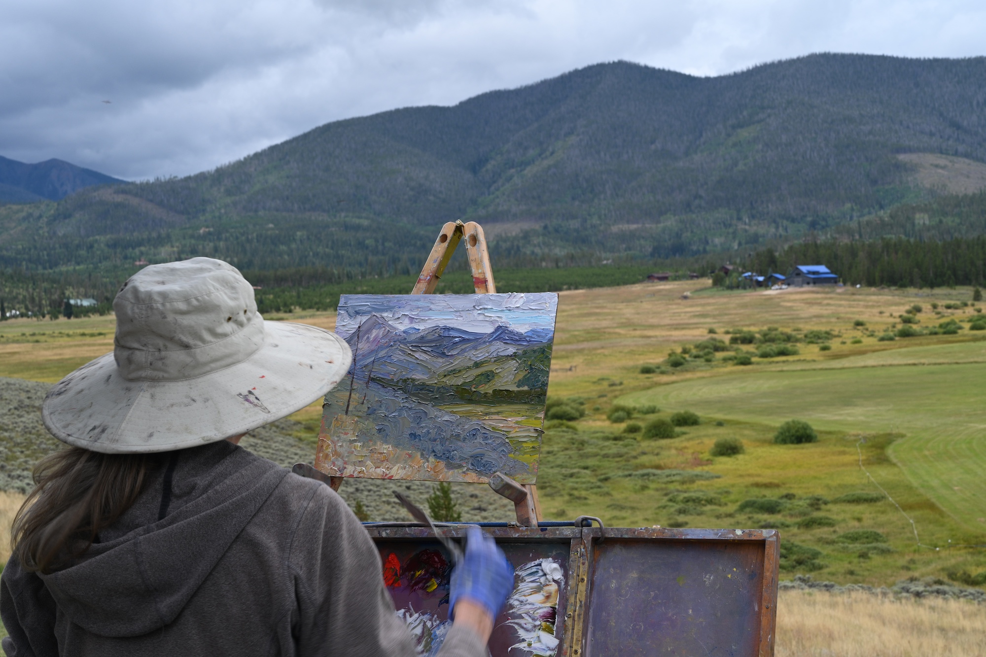 A woman wearing a wide-brimmed hat paints a landscape of rolling hills and distant mountains in Grand County Colorado.