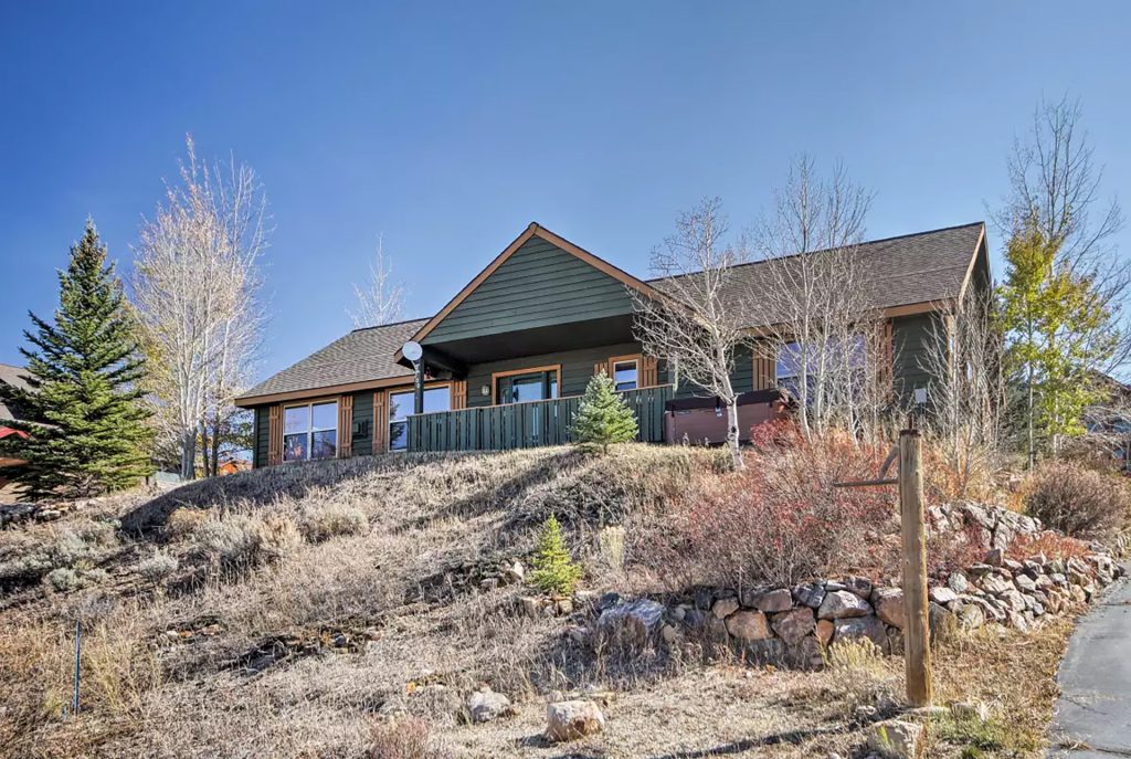 A green, single-story house with a deck and stone wall on a sloped lot with trees in Grand County Colorado.