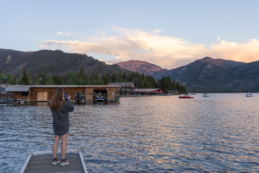 Person standing on a dock taking a photo of the lake, with boats, cabins, and forested mountains in the background at sunset in Grand County.