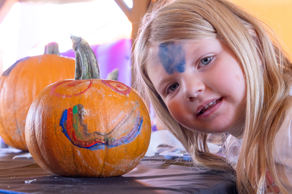 A young girl with face paint smiles next to a decorated pumpkin on a table in Grand County Colorado.