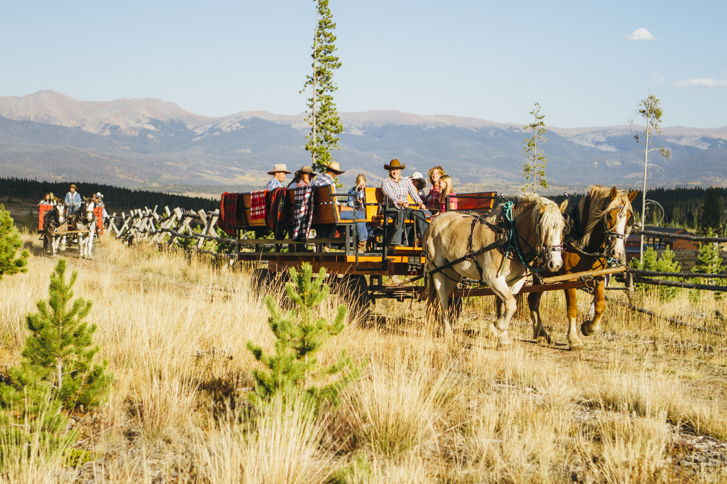 A horse-drawn wagon filled with people travels through a grassy field with mountains in the background in Grand County Colorado.