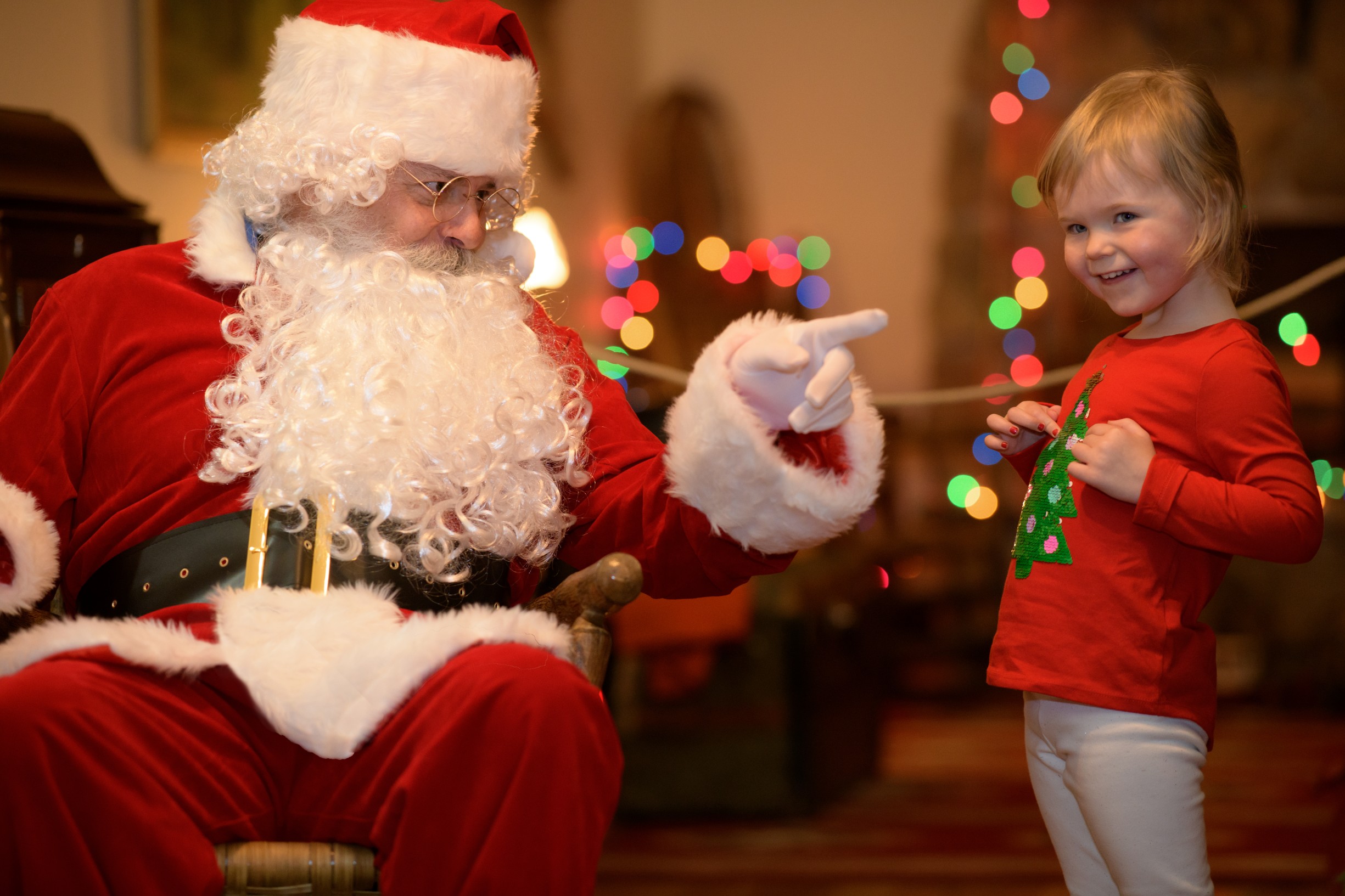Santa Claus in a red suit with a white beard points at a smiling young girl in a Christmas sweater in Grand County Colorado.