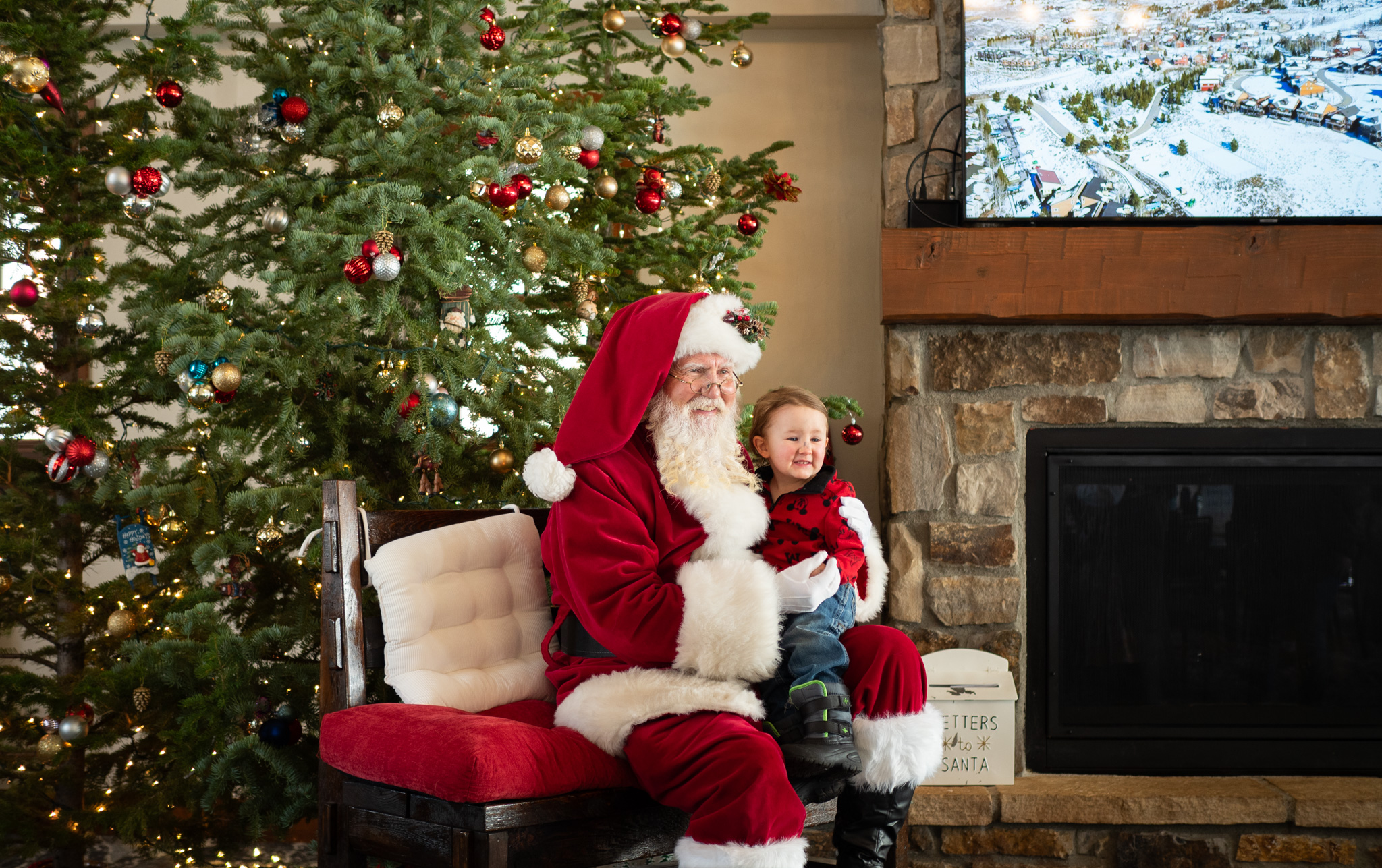 Santa Claus sits with a child in front of a decorated Christmas tree and fireplace in Grand County Colorado.