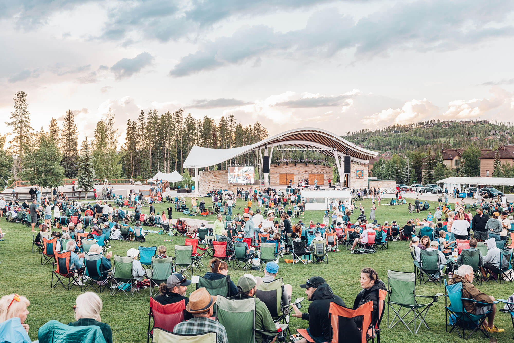 Outdoor concert with a large crowd seated on folding chairs in a grassy area, watching a stage with a curved roof in Grand County Colorado.