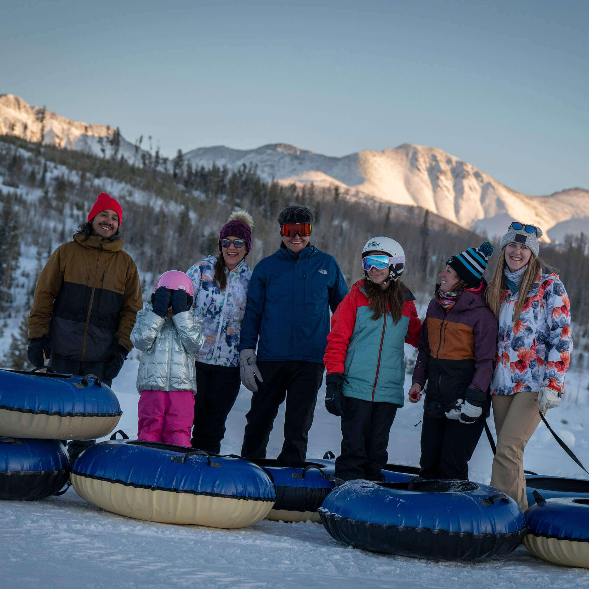 A group of seven people, including a child, are standing in a snowy landscape with blue and yellow inner tubes, ready for a fun day of snow tubing against a backdrop of snow-capped mountains in Grand County Colorado.