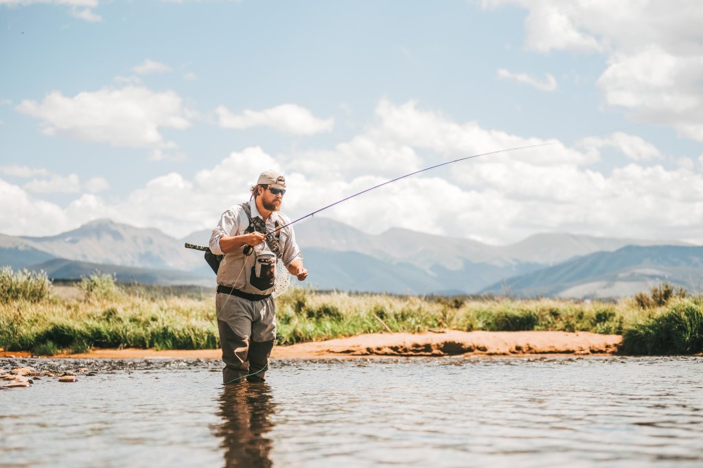 Man fly fishing in shallow river with mountains in the background in Grand County Colorado.