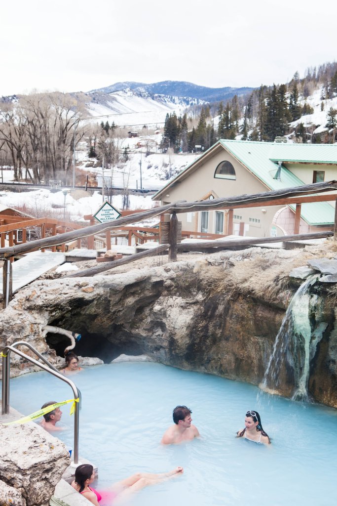 People relax in a natural hot spring pool with blue water surrounded by snow and mountains in Grand County Colorado.