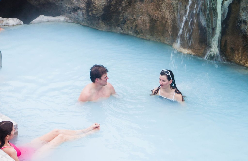 Three people relax in a serene blue hot spring pool surrounded by rocky cliffs, with a waterfall cascading into the water in Grand County Colorado.