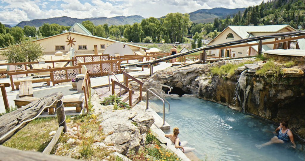 A serene natural hot springs with cascading waterfalls, surrounded by lush greenery and mountain views, offers a tranquil retreat for swimmers and relaxers in Hot Sulphur Springs Spa & Resort in Grand County Colorado.