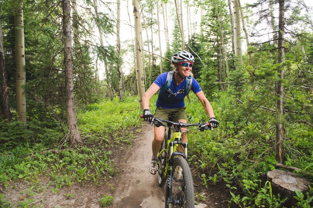 A cyclist in a blue shirt and helmet smiles as they ride a mountain bike along a dirt trail through a lush forest filled with tall trees and green undergrowth at Idlewild Trail System in Grand County Colorado.