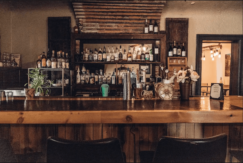 A rustic bar with a wooden counter, shelves stocked with liquor bottles, and hanging light bulbs in Grand County Colorado.