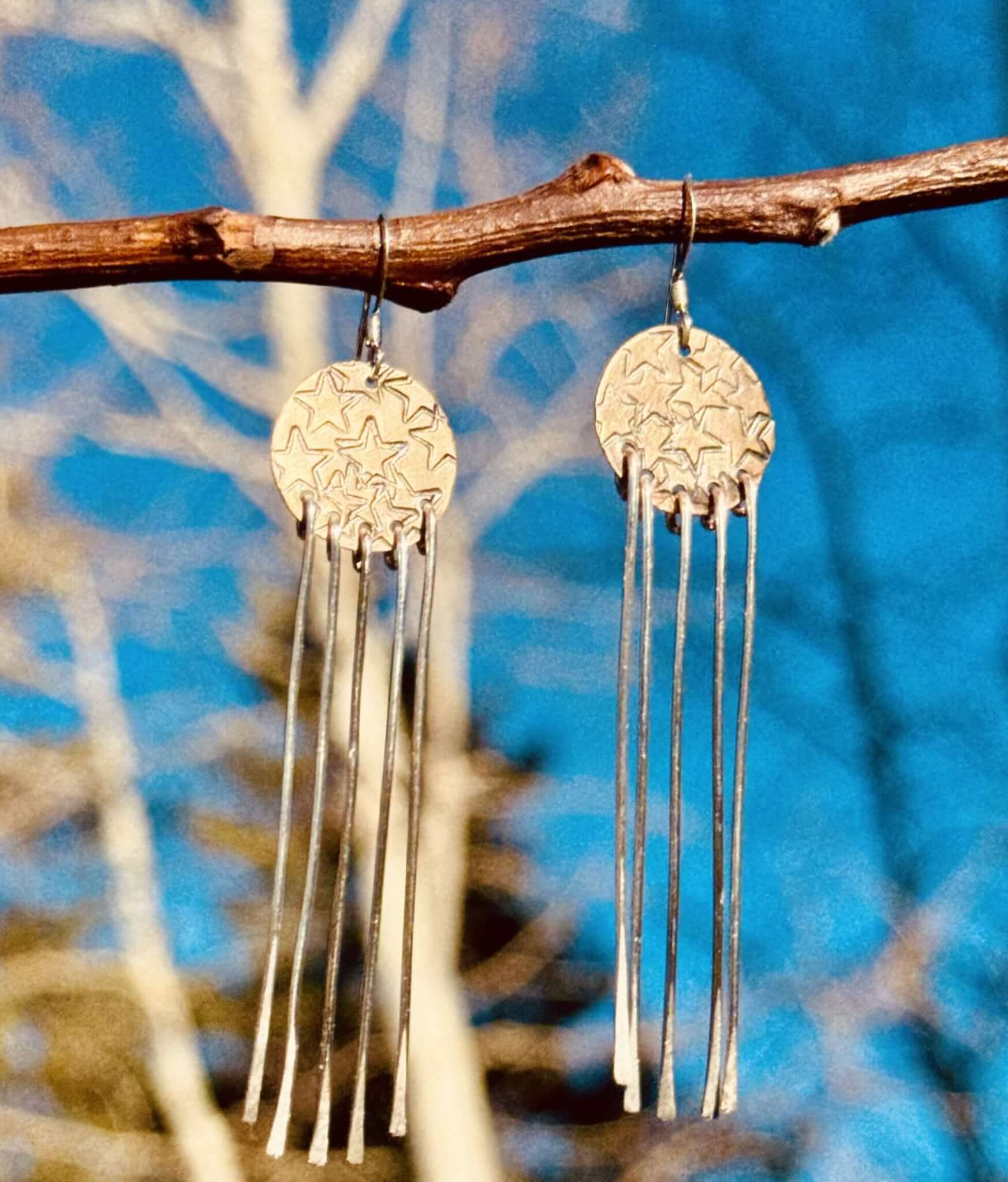 A pair of star-shaped earrings with long, thin metal strands hanging from them, suspended from a tree branch against a clear blue sky in Grand County Colorado.