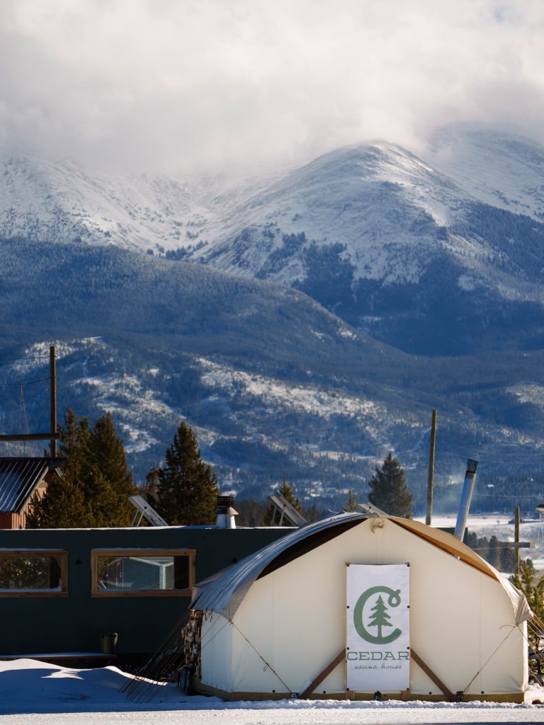 A white tent with a cedar logo stands in the snow, with snow-capped mountains and evergreen trees in the background in Grand County Colorado.
