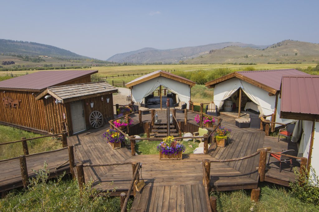 A rustic wooden deck with colorful flower pots and a central fire pit leads to three cozy cabins with canvas walls, nestled in a serene meadow with rolling hills and distant mountains in the background in Grand County Colorado.