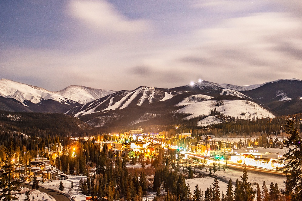 A picturesque mountain town nestled in a snowy valley, illuminated by warm lights against a backdrop of snow-capped peaks under a starry night sky in Grand County Colorado.