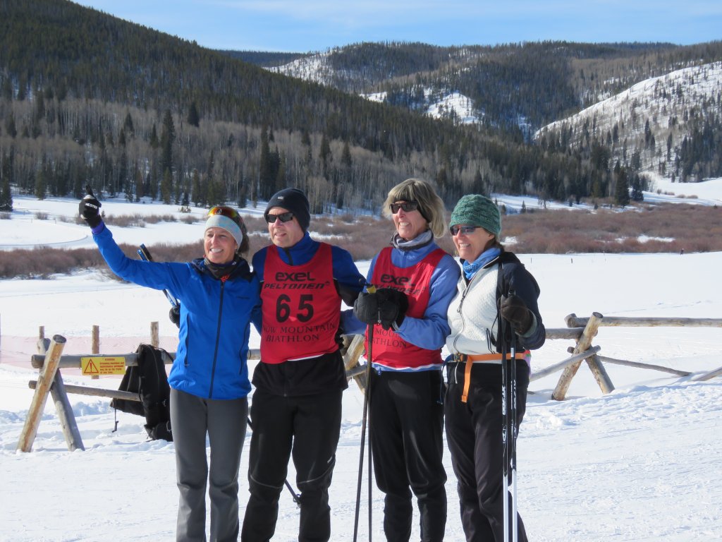 Four skiers, dressed in colorful winter gear and race bibs, celebrate their achievement with a group selfie against a breathtaking backdrop of snow-capped mountains and evergreen forests in Grand County Colorado.