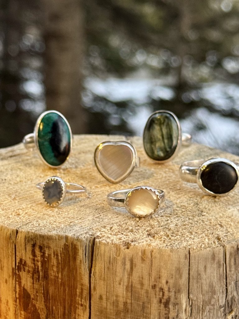 A collection of six unique rings featuring various gemstones and designs, displayed on a rustic wooden stump against a backdrop of a snowy forest in Grand County Colorado.