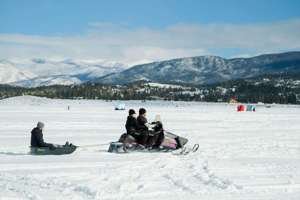 A person on a sled is being pulled by a snowmobile across a vast, snowy landscape with mountains in the distance in Grand County Colorado.